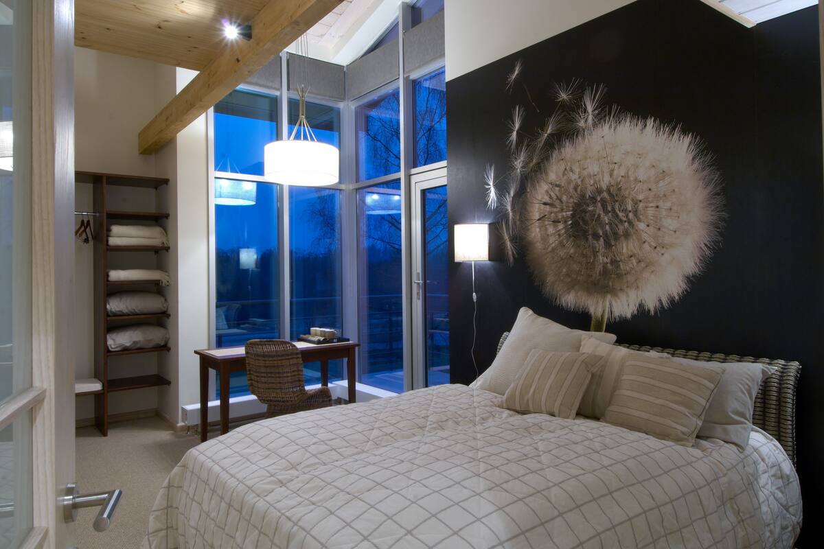 Un dormitorio con una cama grande, una mesita de noche, una silla, una ventana con vistas y una pared decorada con una obra de arte texturizada.