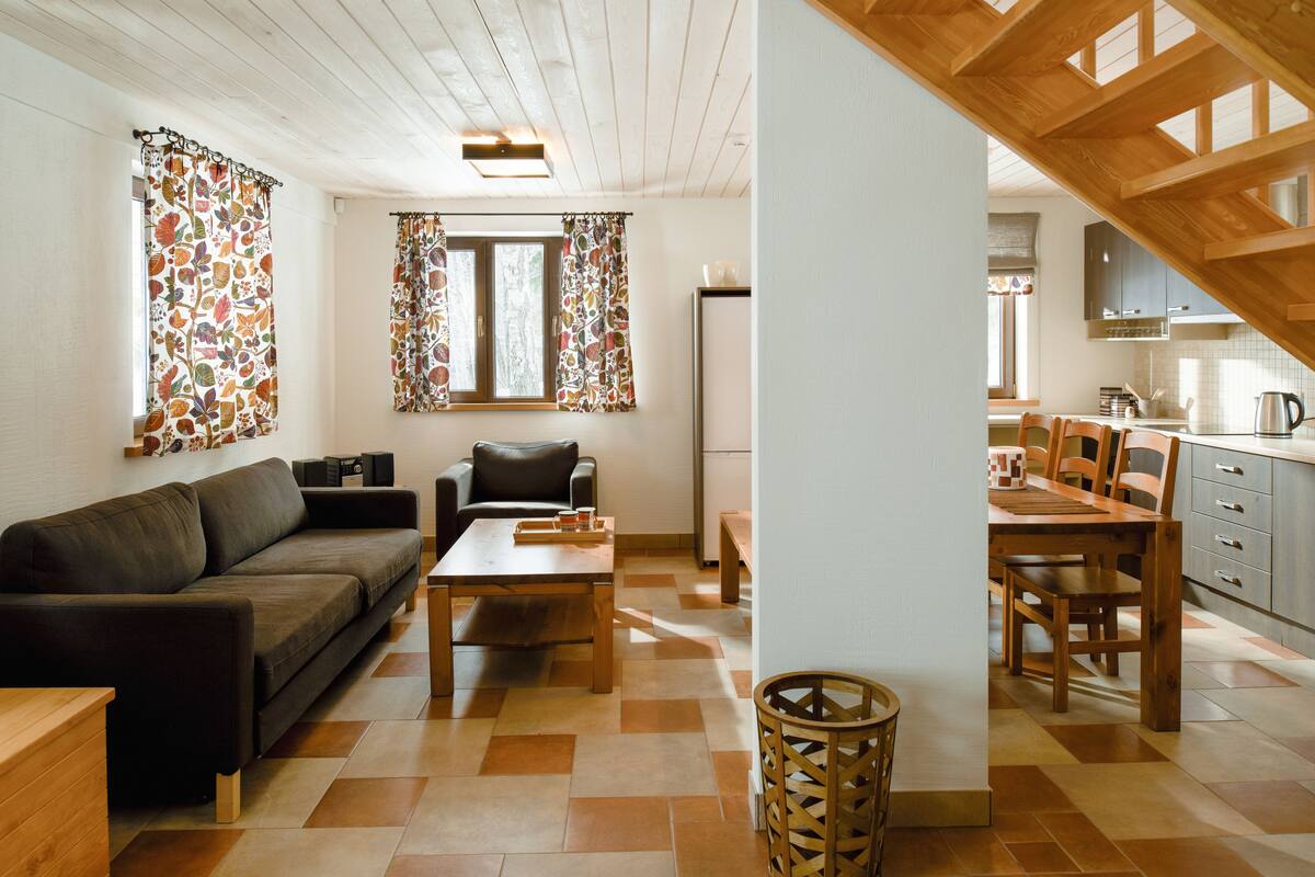 Un salón acogedor con sofá, mesa de centro y zona de comedor con mesa y sillas. Se ve una cocina con armarios y un refrigerador al fondo.