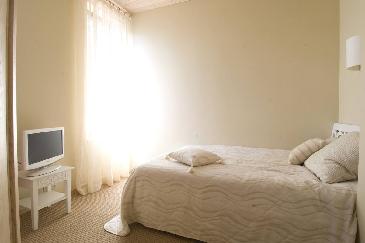 Un dormitorio con una cama, un televisor sobre una mesita y una ventana con cortinas.