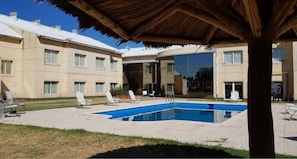 Uma área de piscina com espreguiçadeiras, um espaço de estar coberto e um edifício com várias janelas e uma porta de vidro.