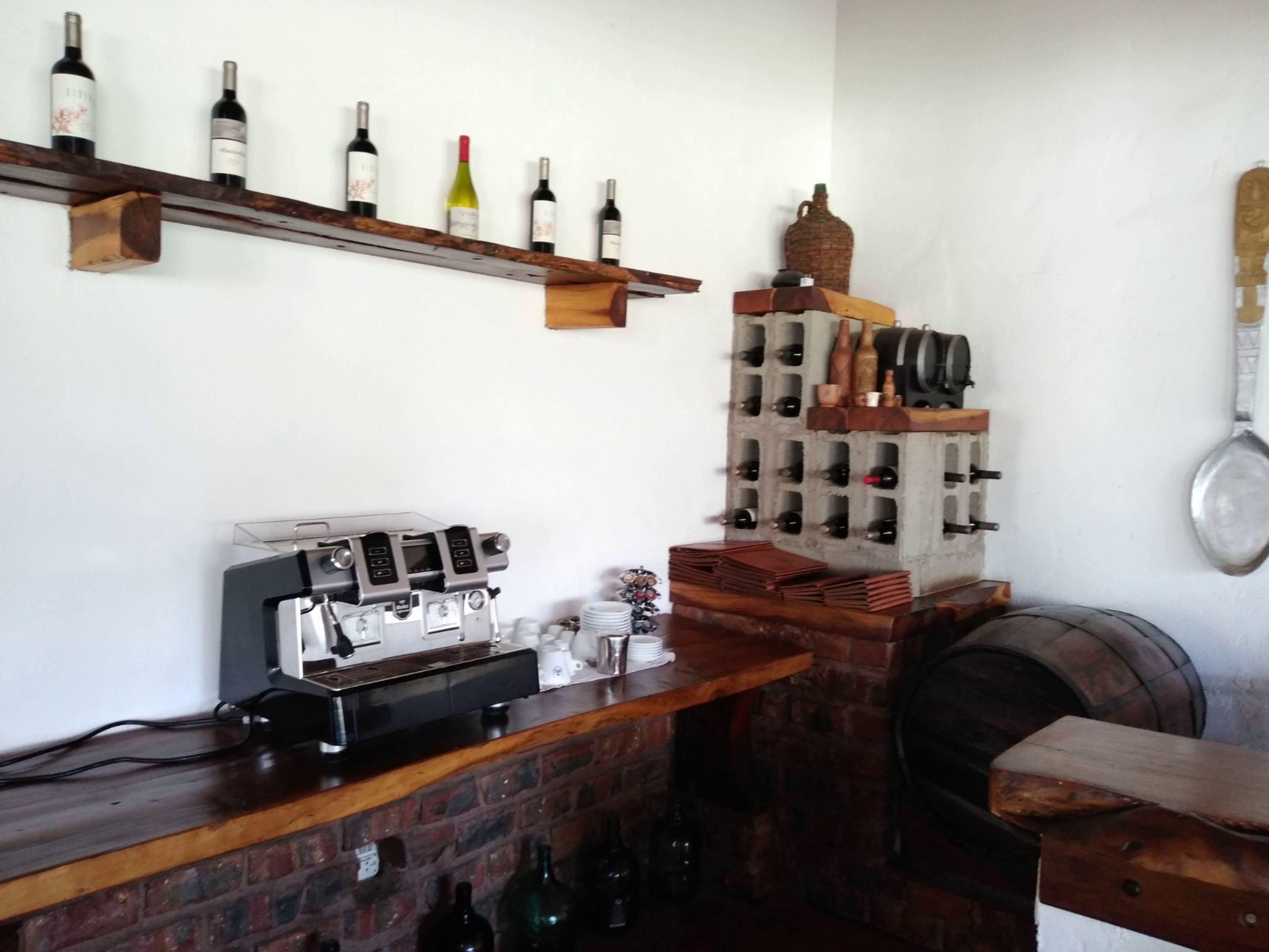 Zona de bar con pared de ladrillo, estantes de madera con botellas de vino, una máquina de café y una pila de posavasos.