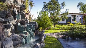 A landscaped garden with a rock waterfall, a paved pathway, and a building with a white facade and green shutters. - Casa Hacienda Nasca Oasis (Nazca)
