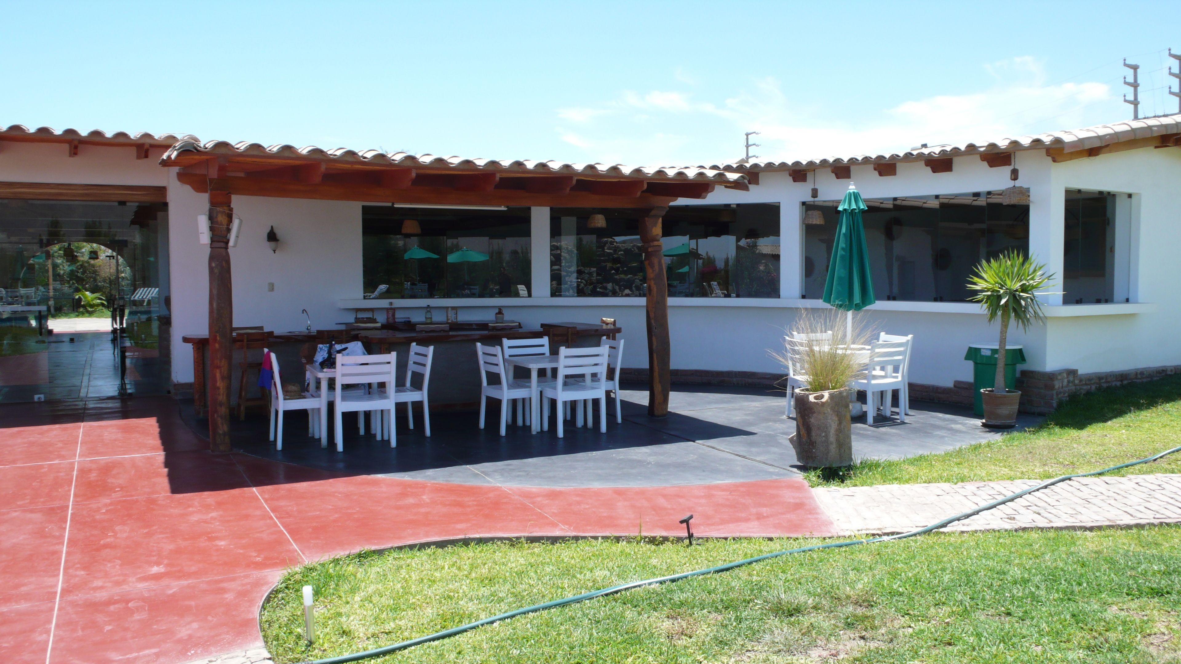 A patio with a wooden pergola, white chairs, and a table. There is a green umbrella and a potted plant.