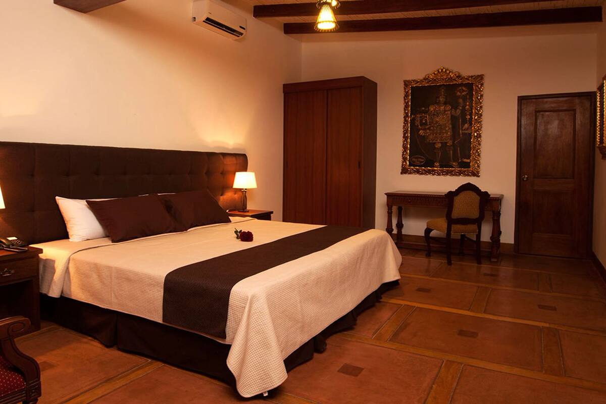 Una habitación de hotel con una cama grande, dos lámparas de noche, un cabecero de madera, un escritorio de madera con una silla y un cuadro en la pared.