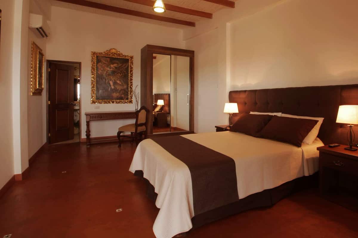 Habitación de hotel con una cama grande, dos lámparas de noche, una mesita de noche de madera, una silla, un escritorio y un cuadro en la pared.
