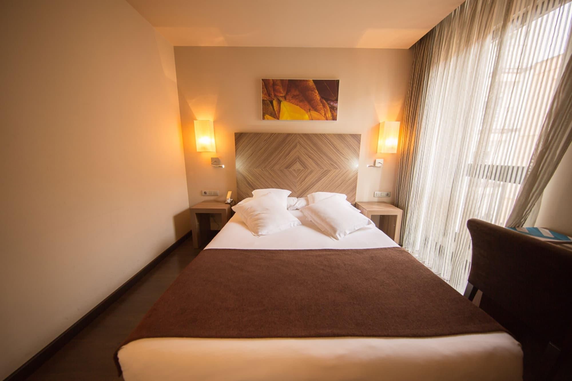 A hotel room with a bed, two bedside tables, a headboard, a painting, and a window with curtains.