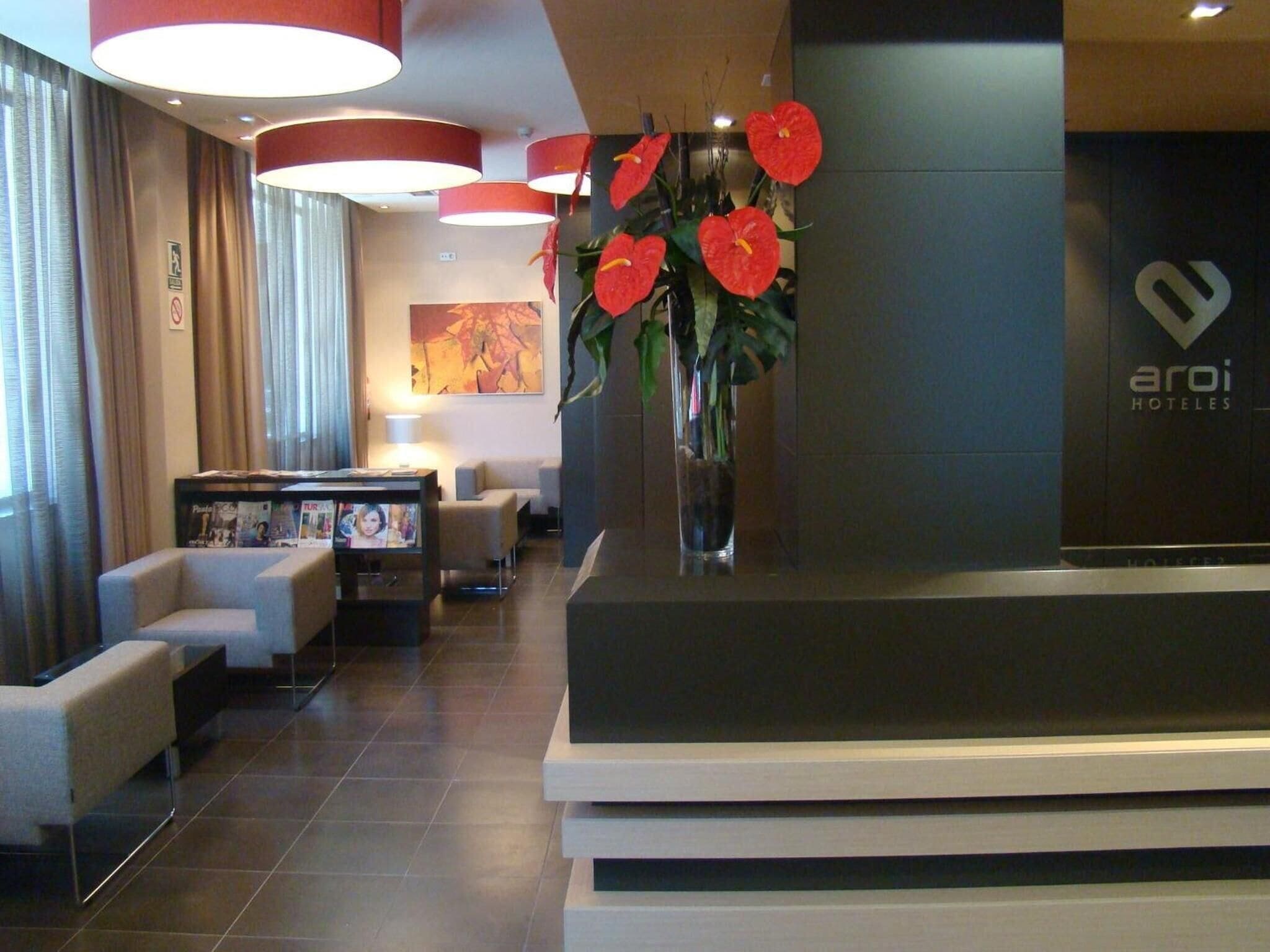 Zona de recepción de un hotel con diseño moderno, que incluye un mostrador de recepción, un área de descanso con sofás y un jarrón grande con flores rojas.