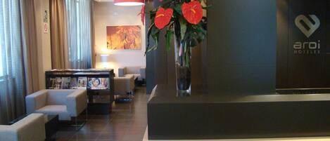 Zona de recepción de un hotel con diseño moderno, que incluye un mostrador de recepción, un área de descanso con sofás y un jarrón grande con flores rojas.
