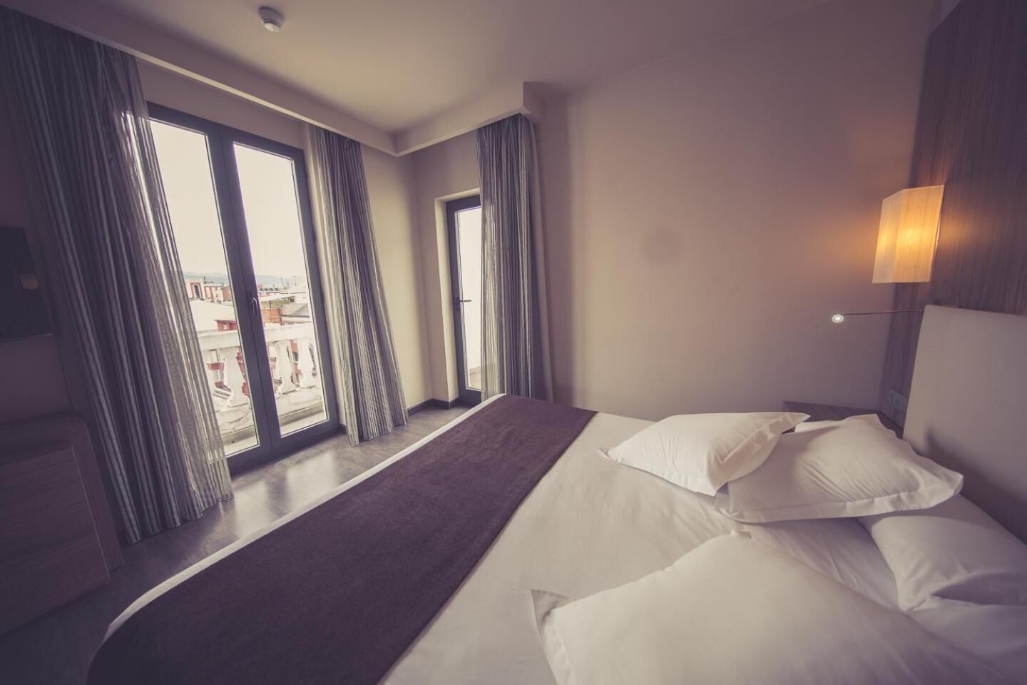 Une chambre d’hôtel avec un grand lit, deux fenêtres avec des rideaux, une vue sur des immeubles et une lampe fixée au mur.