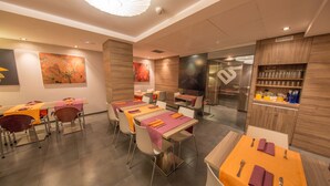 A modern restaurant interior with multiple dining tables, chairs, and a kitchen area visible through glass doors.
