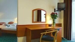 Habitación de hotel con cama, un escritorio de madera con silla, espejo, televisor y un jarrón con flores.