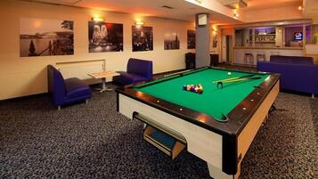 A pool table with cue balls, two blue chairs, and a bar area with a neon sign.