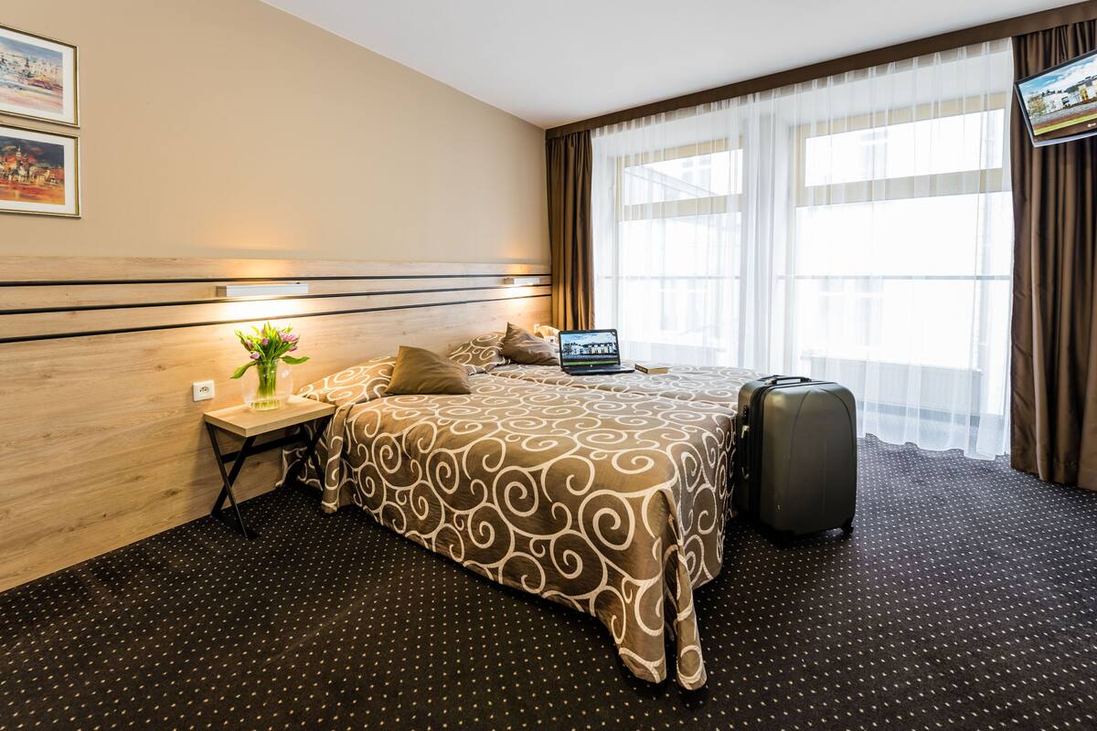 A hotel room with a bed, a desk with a laptop, a suitcase, and a window with curtains.