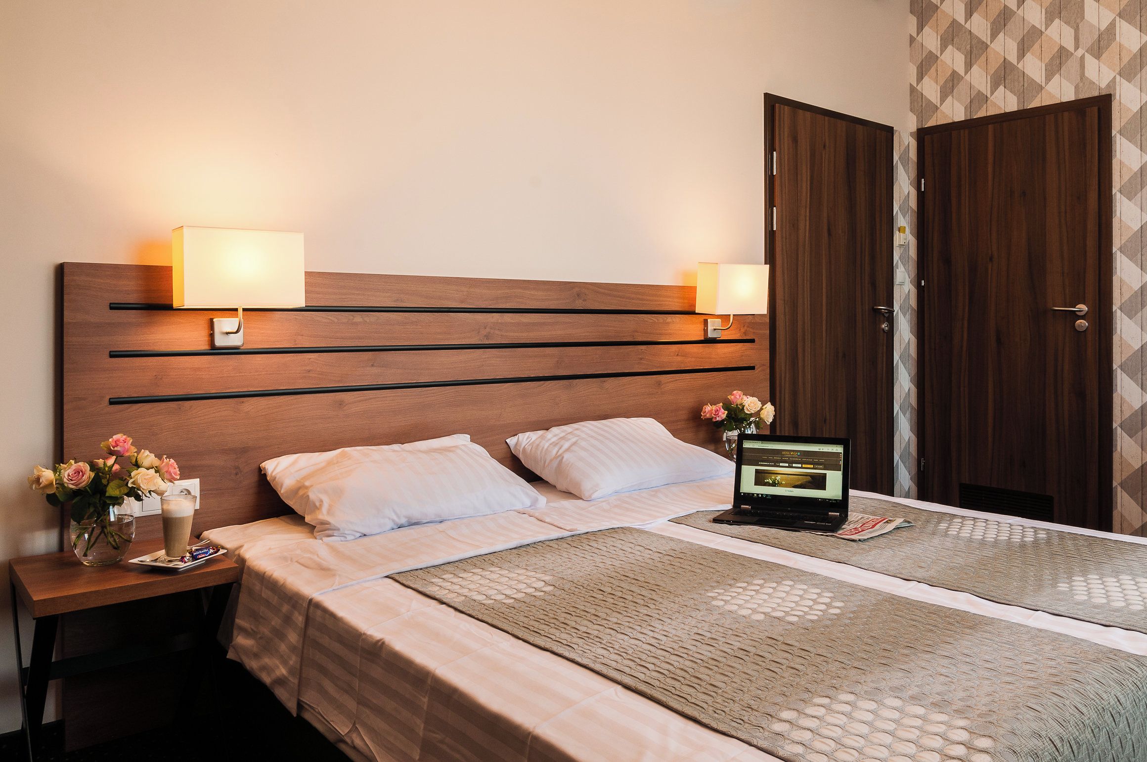 A hotel room with a wooden headboard, two bedside lamps, a laptop on the bed, a vase with flowers, and a minibar.