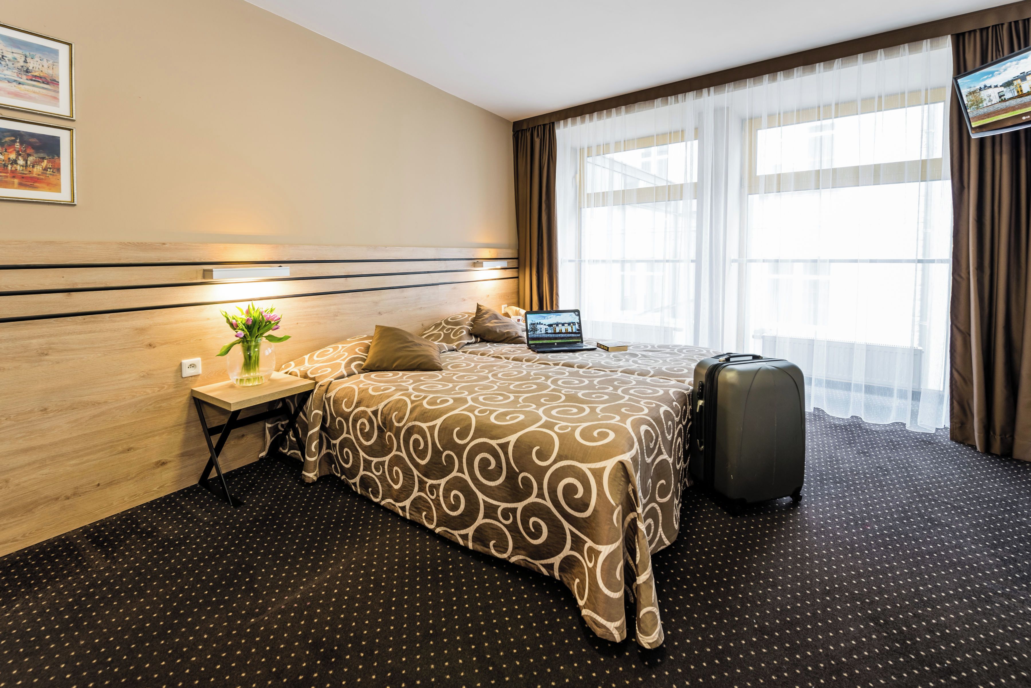 A hotel room with a bed, a desk with a laptop, a suitcase, and a window with curtains.