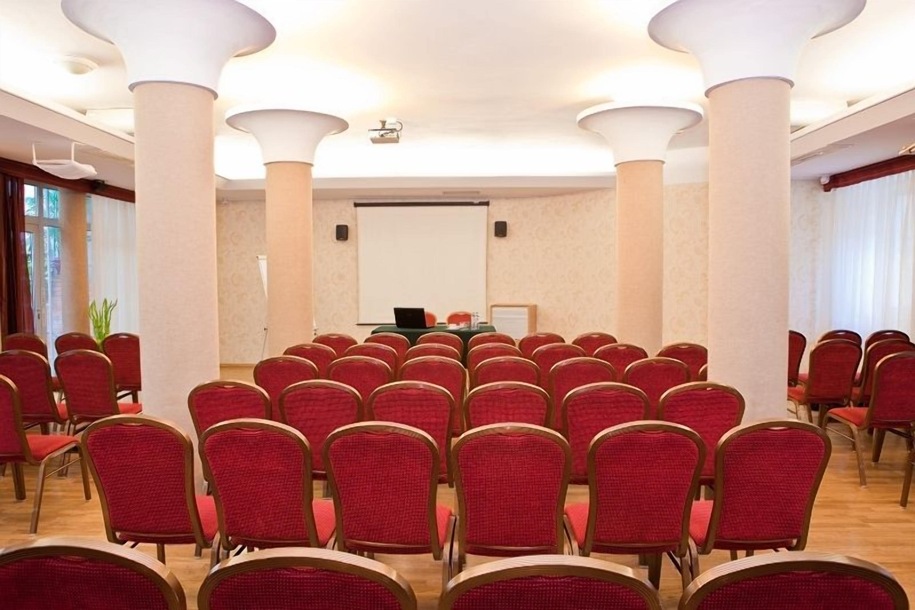 Een vergaderzaal met rijen rode stoelen, een projectiescherm en een presentatietafel.