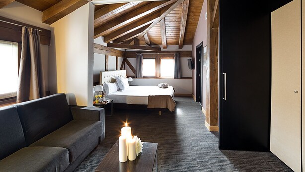 Un dormitorio con cama, mesita de noche y velador. Hay una ventana con cortinas, una puerta y un techo de madera.