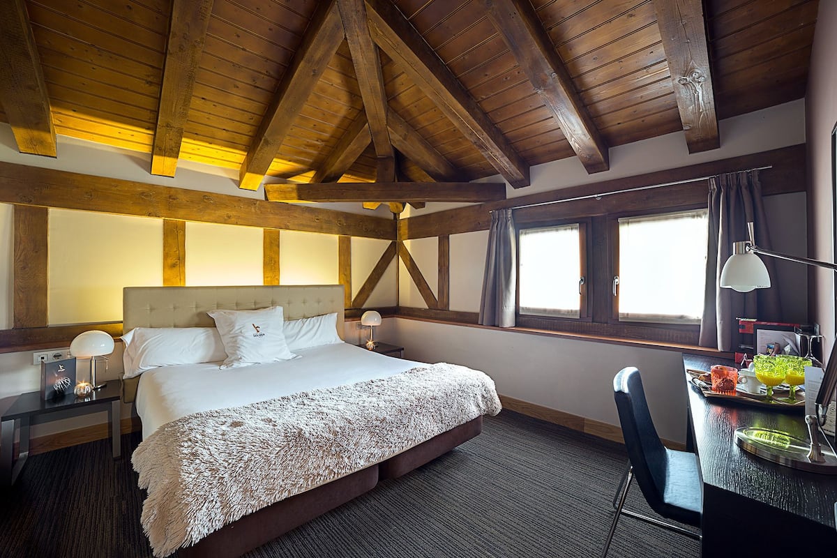 A bedroom with a large bed, wooden beams, a desk with a lamp, and a window with curtains.
