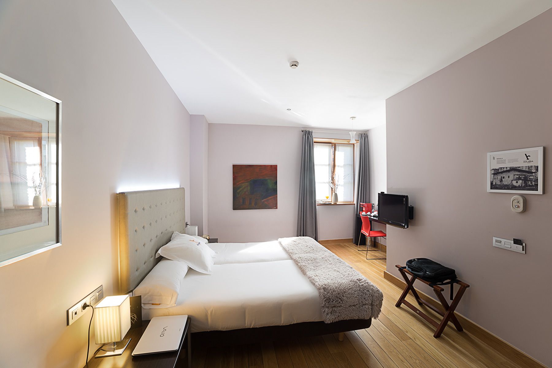 A hotel room with a bed, bedside table, lamp, and a desk with a chair. There is a window with curtains, a painting on the wall, and a television mounted on the wall.