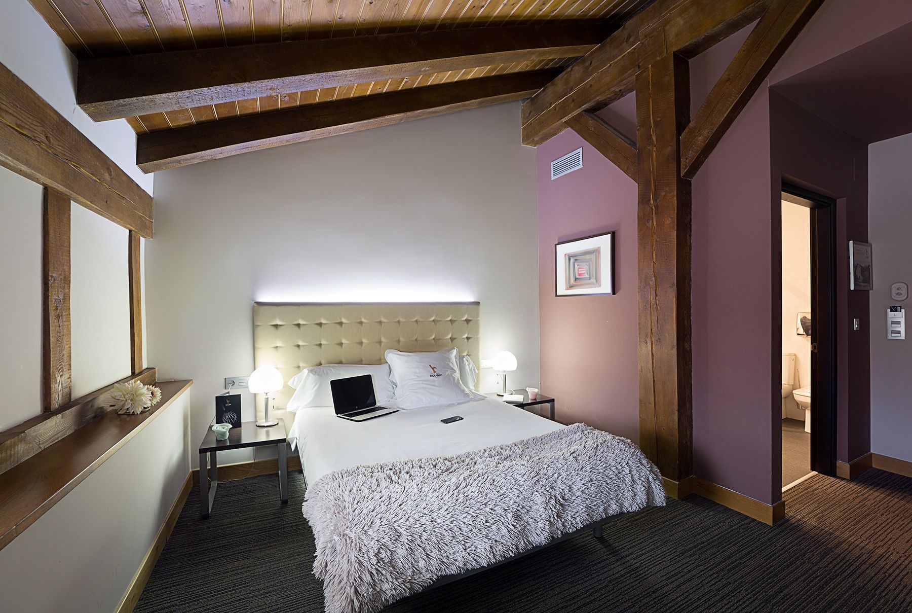 Een hotelkamer met een groot bed, houten balken, een nachtkastje met lamp en een deur naar een badkamer.