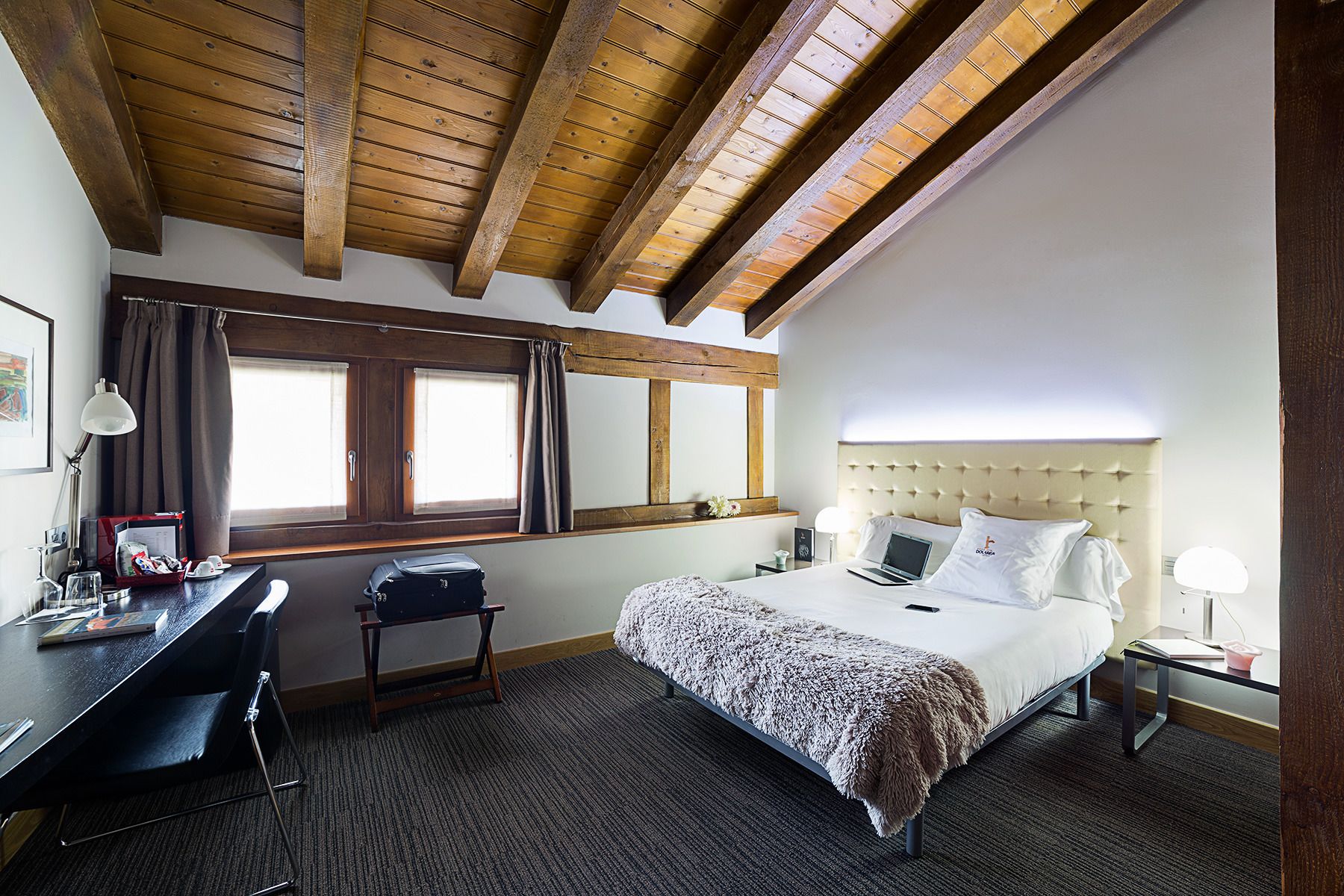 A hotel room with a wooden ceiling, a bed with a tufted headboard, a desk with a chair, and a window with curtains.