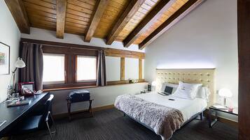 A hotel room with a wooden ceiling, a bed with a tufted headboard, a desk with a chair, and a window with curtains.