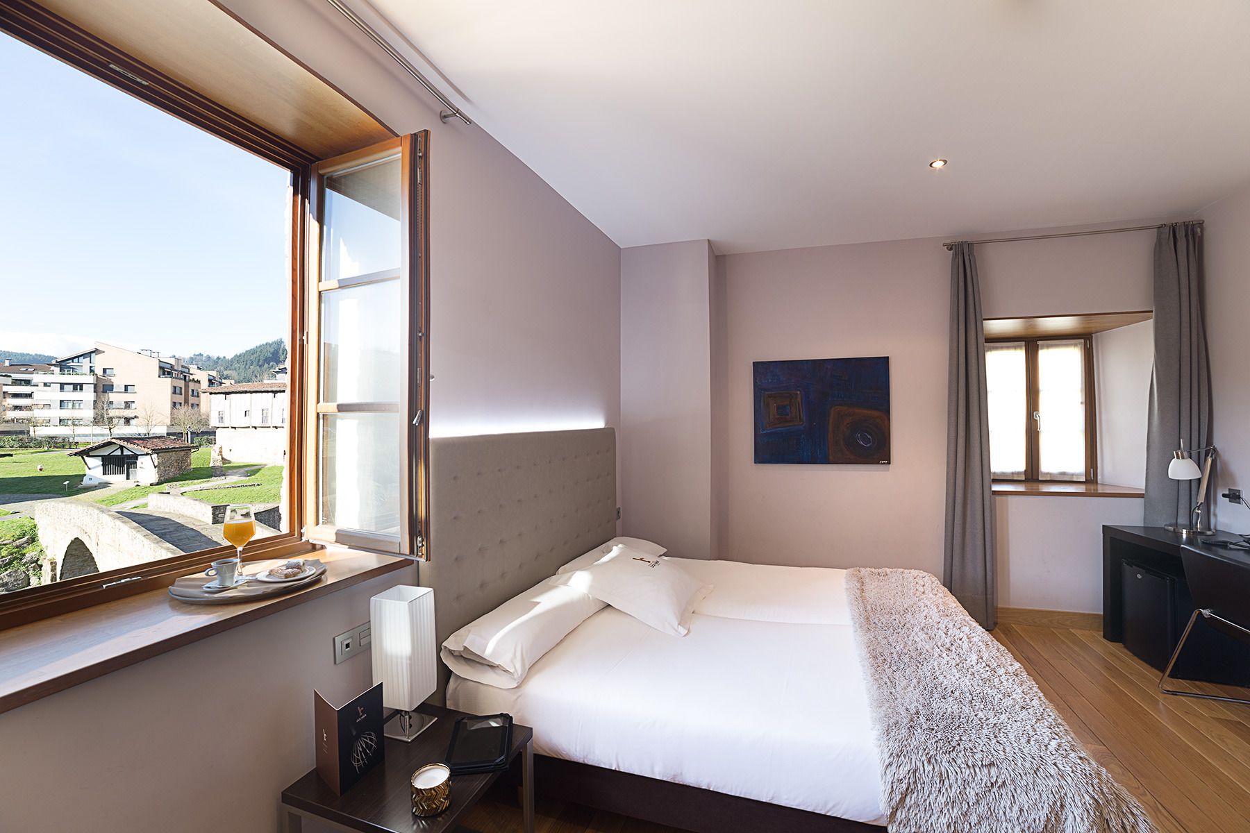 Habitación de hotel con cama, mesita de noche, lámpara y vistas al exterior.