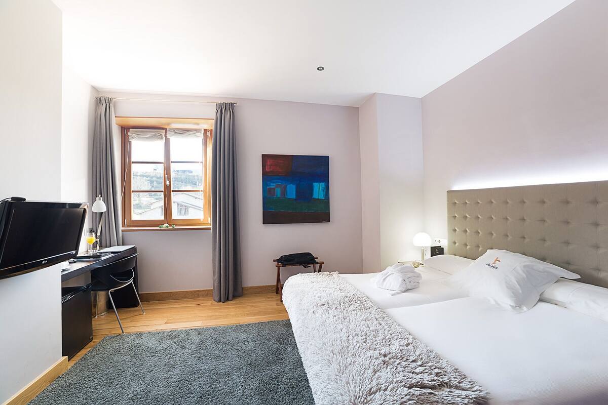 A hotel room with a bed, a desk with a computer, a window with curtains, a painting on the wall, and a nightstand with a lamp.
