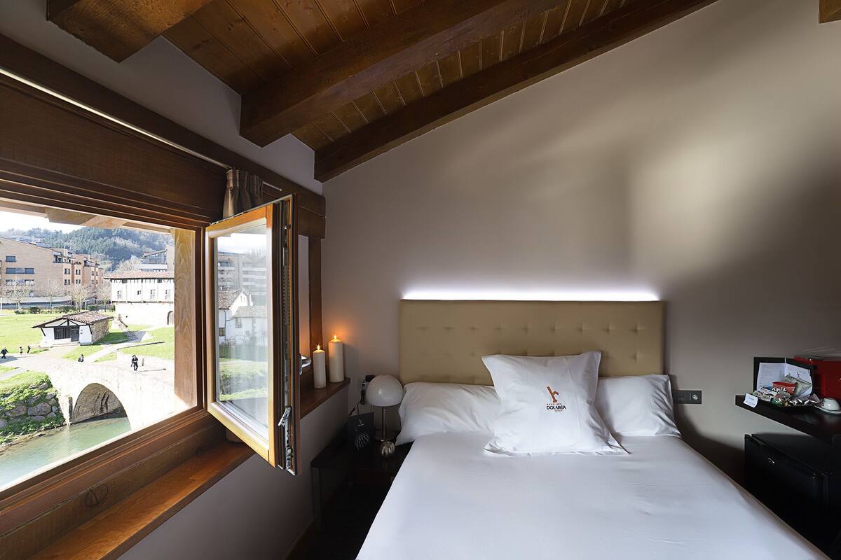 A hotel room with a bed, a nightstand, a lamp, and a window with a view of a bridge and buildings.