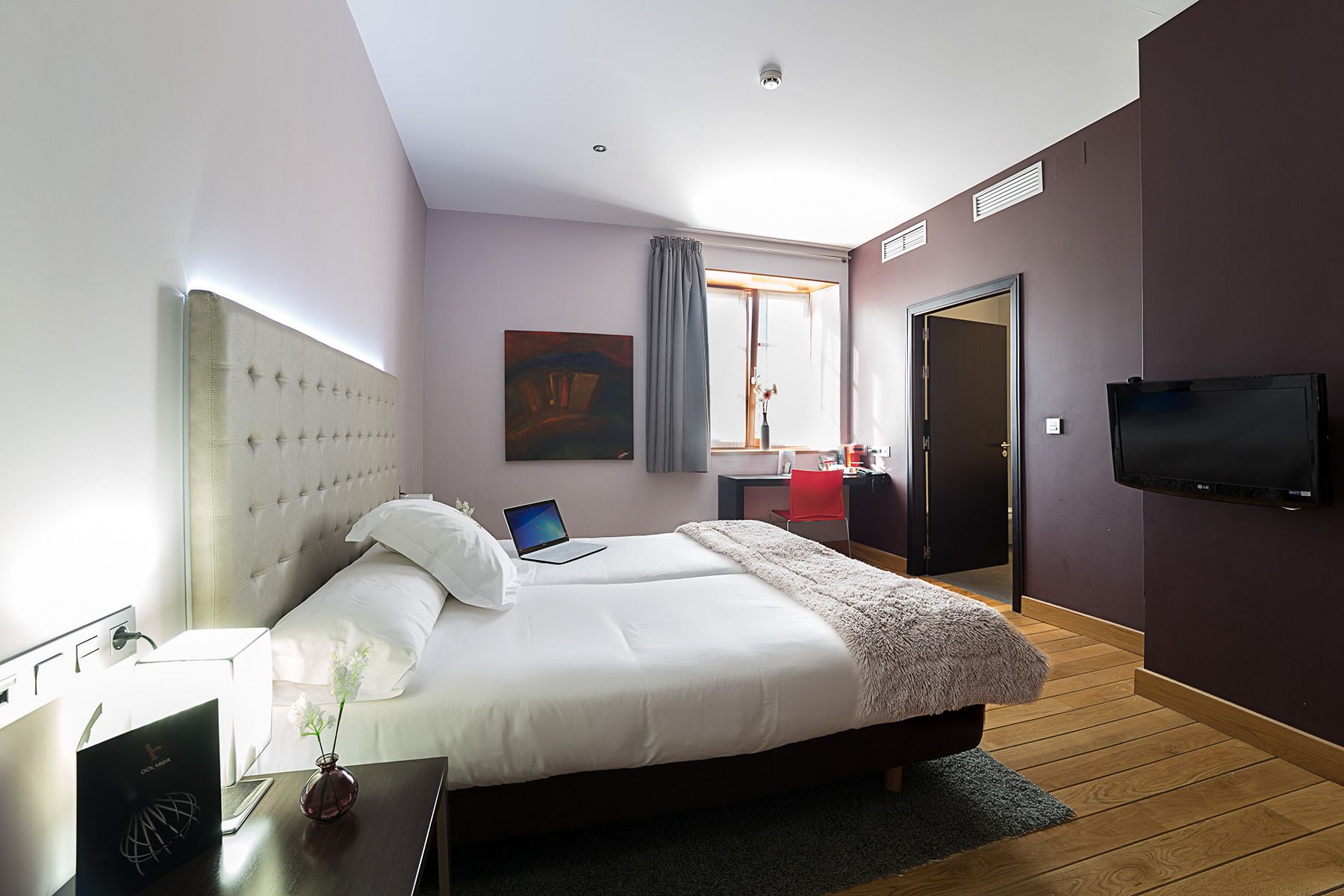 Ein modernes Hotelzimmer mit einem großen Bett, einem Schreibtisch mit einem Laptop, einem Fernseher und einem Fenster mit Vorhängen.