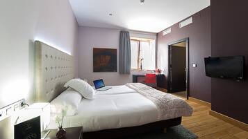 Ein modernes Hotelzimmer mit einem großen Bett, einem Schreibtisch mit einem Laptop, einem Fernseher und einem Fenster mit Vorhängen.