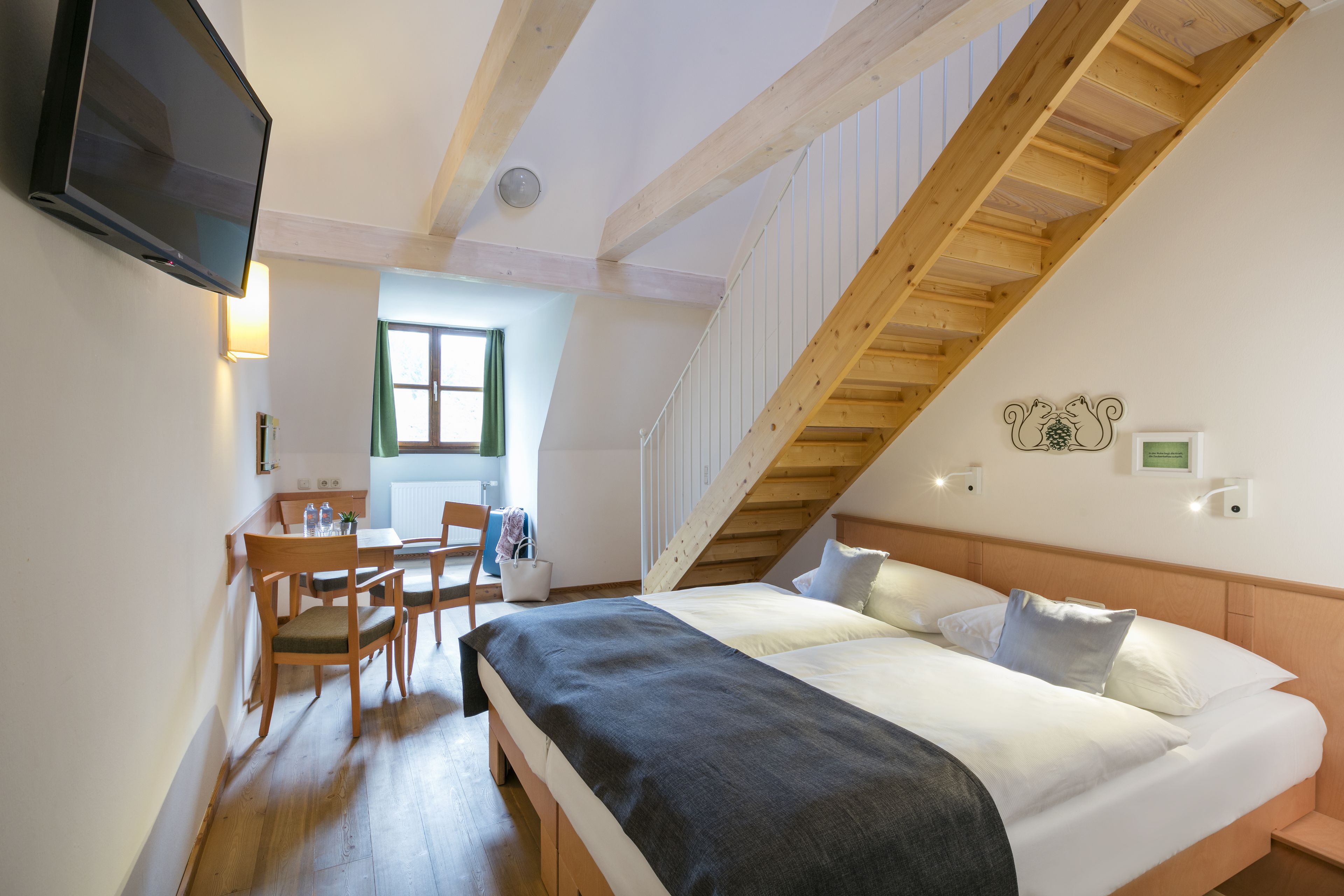 Een slaapkamer met een bed, houten balken in het plafond, een televisie, een bureau met stoel en een raam.