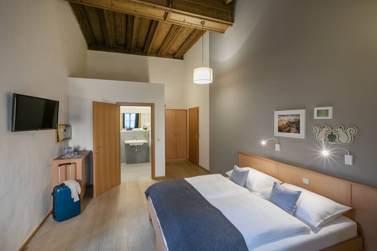 Hotelkamer met een bed, een televisie, een bureau met stoel, een koffer en een zichtbare badkamer door een open deur.