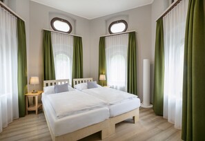 A double bed with white bedding, two bedside tables with lamps, green curtains, and a wooden floor.