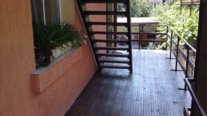 Um deck de madeira com guarda-corpo de metal, um vaso de planta e uma escadaria que leva a um andar superior.