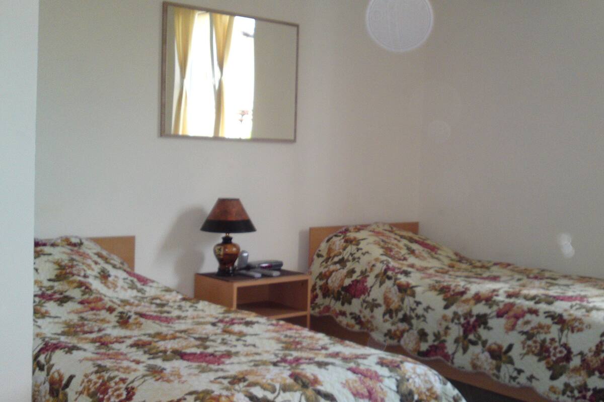 Una habitación con dos camas individuales, cada una con ropa de cama floral. Hay una mesita de noche con una lámpara entre las camas. Un espejo cuelga de la pared sobre las camas.
