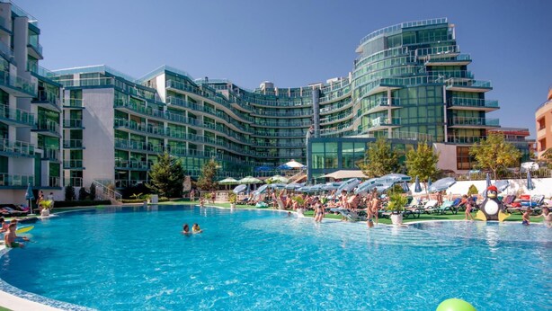 Una piscina rodeada de edificios modernos con balcones, cielo despejado y sombrillas.