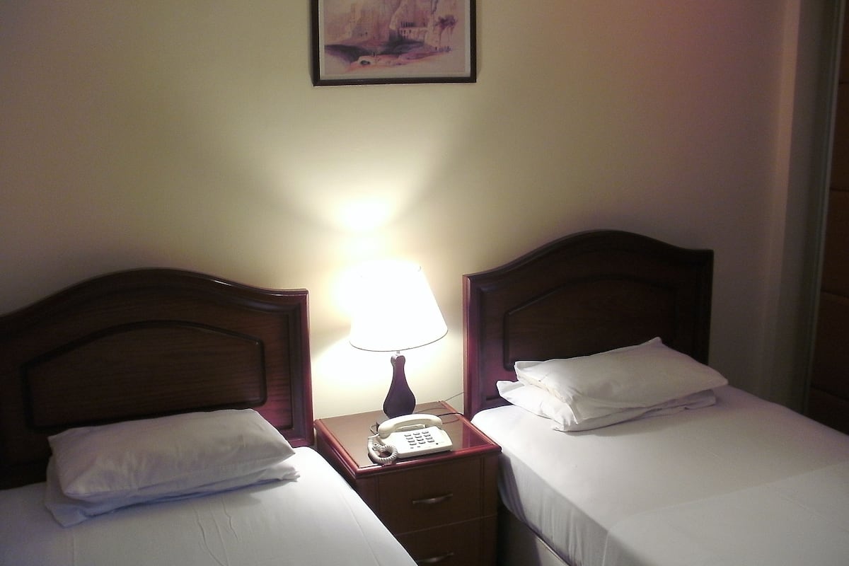 A hotel room with two single beds, a nightstand with a lamp and a telephone, and a framed picture on the wall.