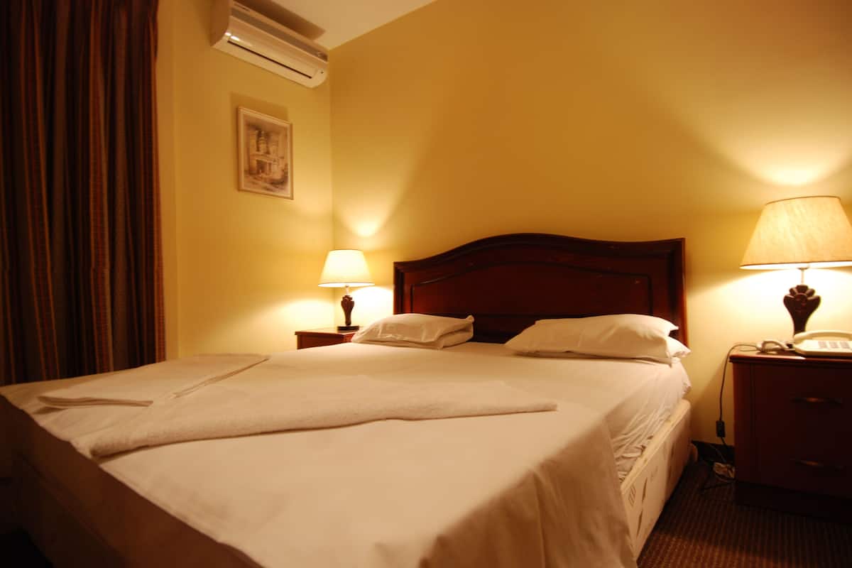 A hotel room with a large bed, two bedside lamps, a wooden headboard, a nightstand, and an air conditioning unit on the wall.