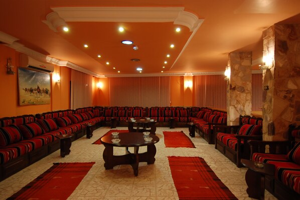 A large, well-lit room with a central seating area featuring multiple sofas and tables, surrounded by stone walls and red carpets.