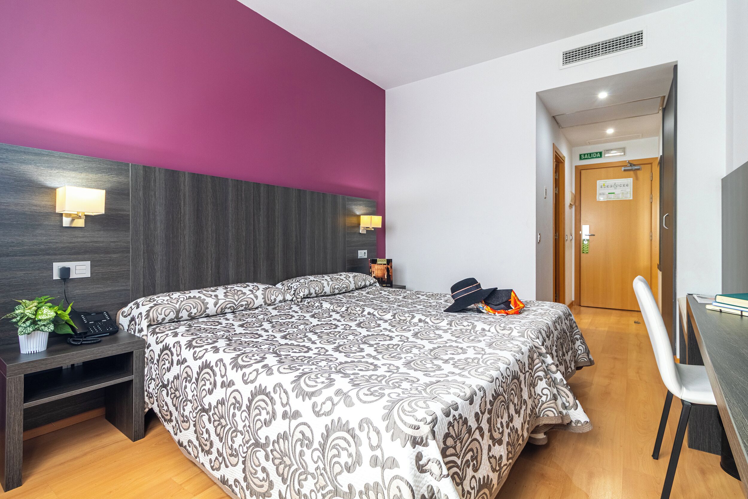 Une chambre d’hôtel avec un grand lit, des tables de chevet, un bureau avec une chaise, une plante en pot et une porte portant l’inscription « SORTIE ».