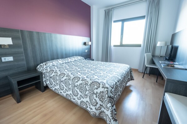 Habitación de hotel con cama, escritorio, silla, ventana con cortinas y televisión.