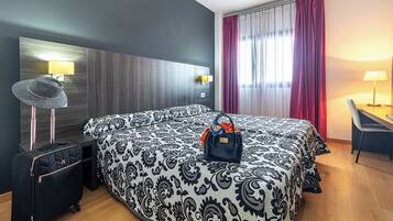 Camera d'albergo con un letto grande, una scrivania, una sedia, una valigia e un cappello sul comodino.