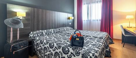Una habitación de hotel con una cama grande, un escritorio, una silla, una maleta y un sombrero sobre la mesita de noche.