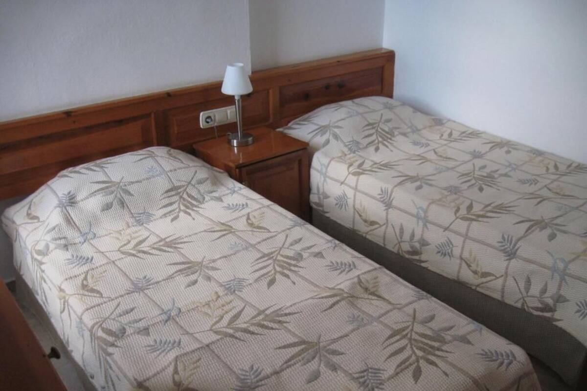 A room with two single beds with leaf-patterned bedding, a nightstand with a lamp, and a wooden headboard.