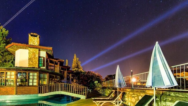 Une vue nocturne d’un complexe hôtelier avec une piscine, des chaises longues et un bâtiment en pierre doté d’une cheminée.