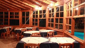 Un espace repas avec des tables et des chaises en bois, de grandes fenêtres et une piscine visible à travers ces fenêtres.