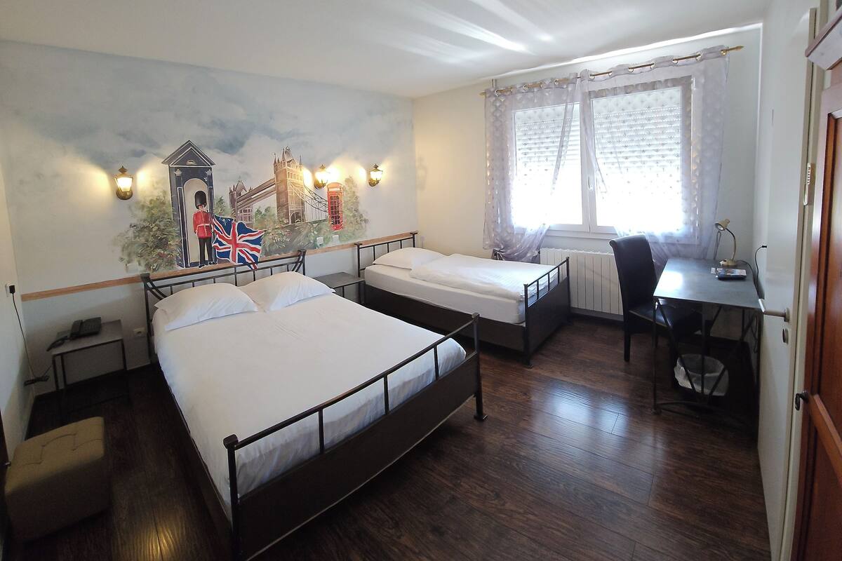 A hotel room with two single beds, a desk, a chair, a radiator, and a mural of a cityscape.