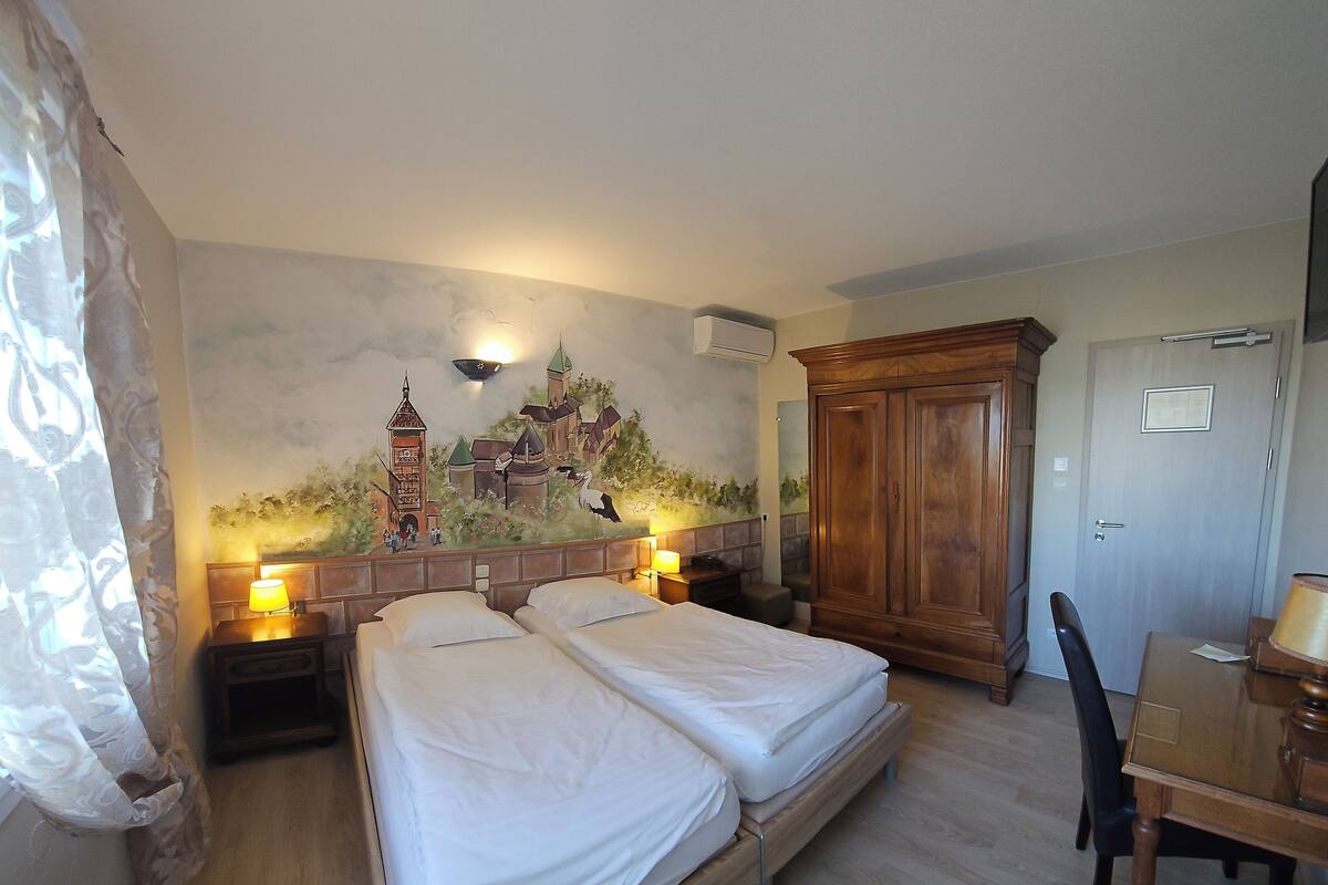A hotel room with two beds, a wooden wardrobe, a desk with a chair, and a mural on the wall.
