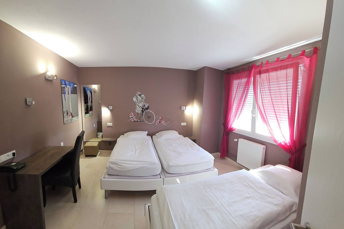 A hotel room with two single beds, a desk with a chair, a small table, and a window with pink curtains.